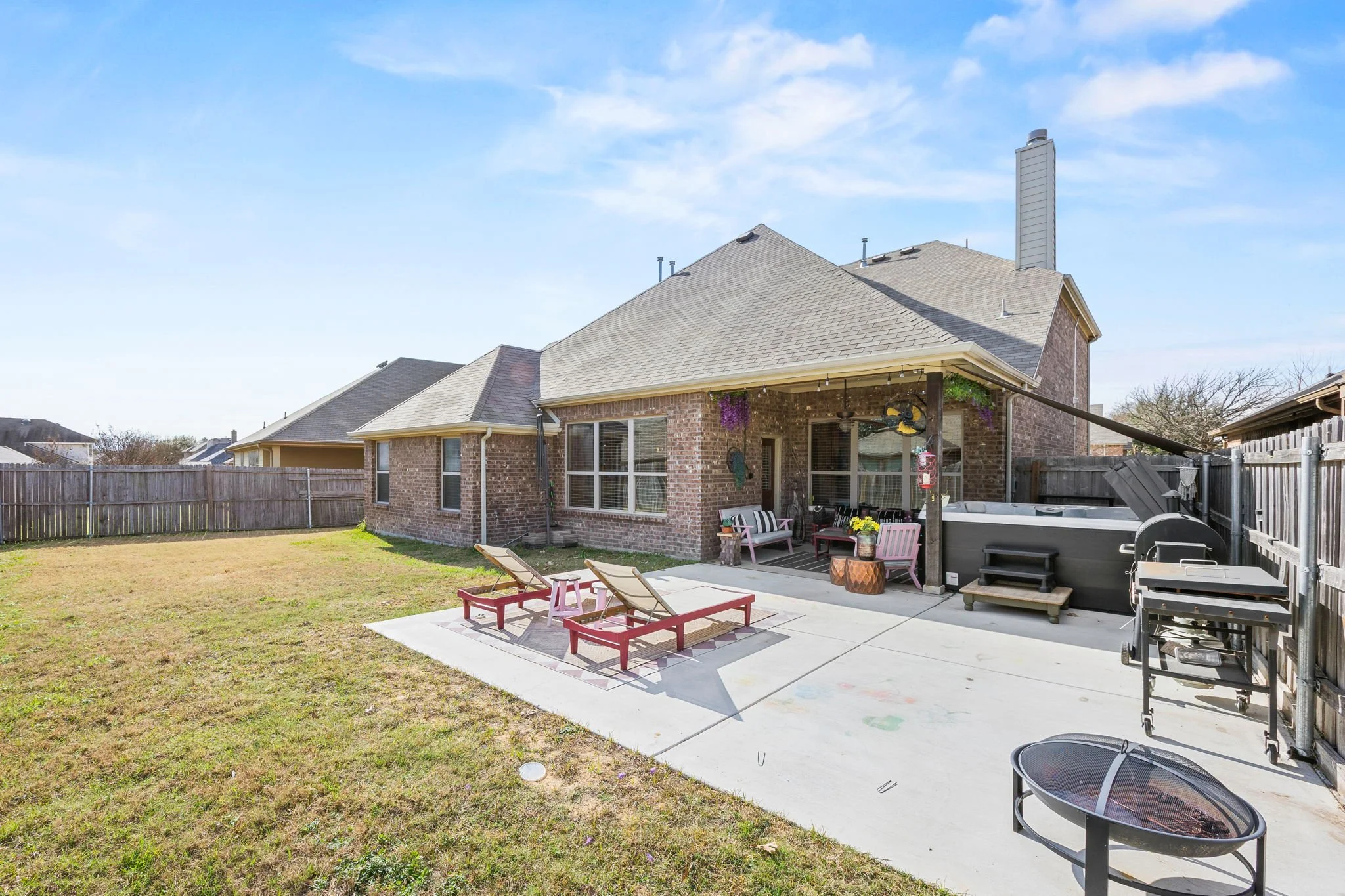 Backyard with a brick house, outdoor grill, lounging chairs, porch furniture, and a fenced yard under a blue sky.