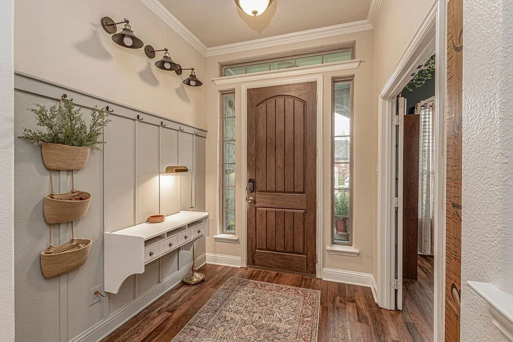 Entryway with wooden front door, side windows, a rug, wall-mounted light, and a wall organizer with potted plants.