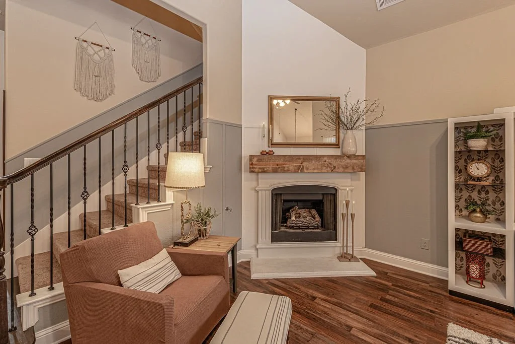 Cozy living room with beige sofa, wooden side table and lamp, fireplace with mirror and dried flowers, decorative bookshelf, and staircase with textured wall hangings.