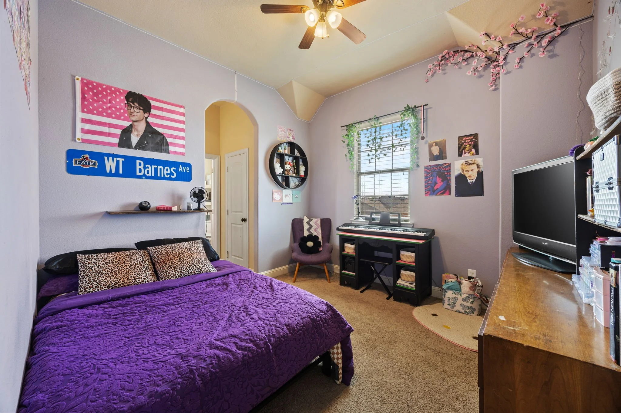 A bedroom with a purple bedspread, leopard print pillows, and a black headboard. A wall-mounted shelf with decorations is above the bed. There is a sign reading "WT Barnes Ave" and a poster of a man with glasses and a flag background. A window with h
