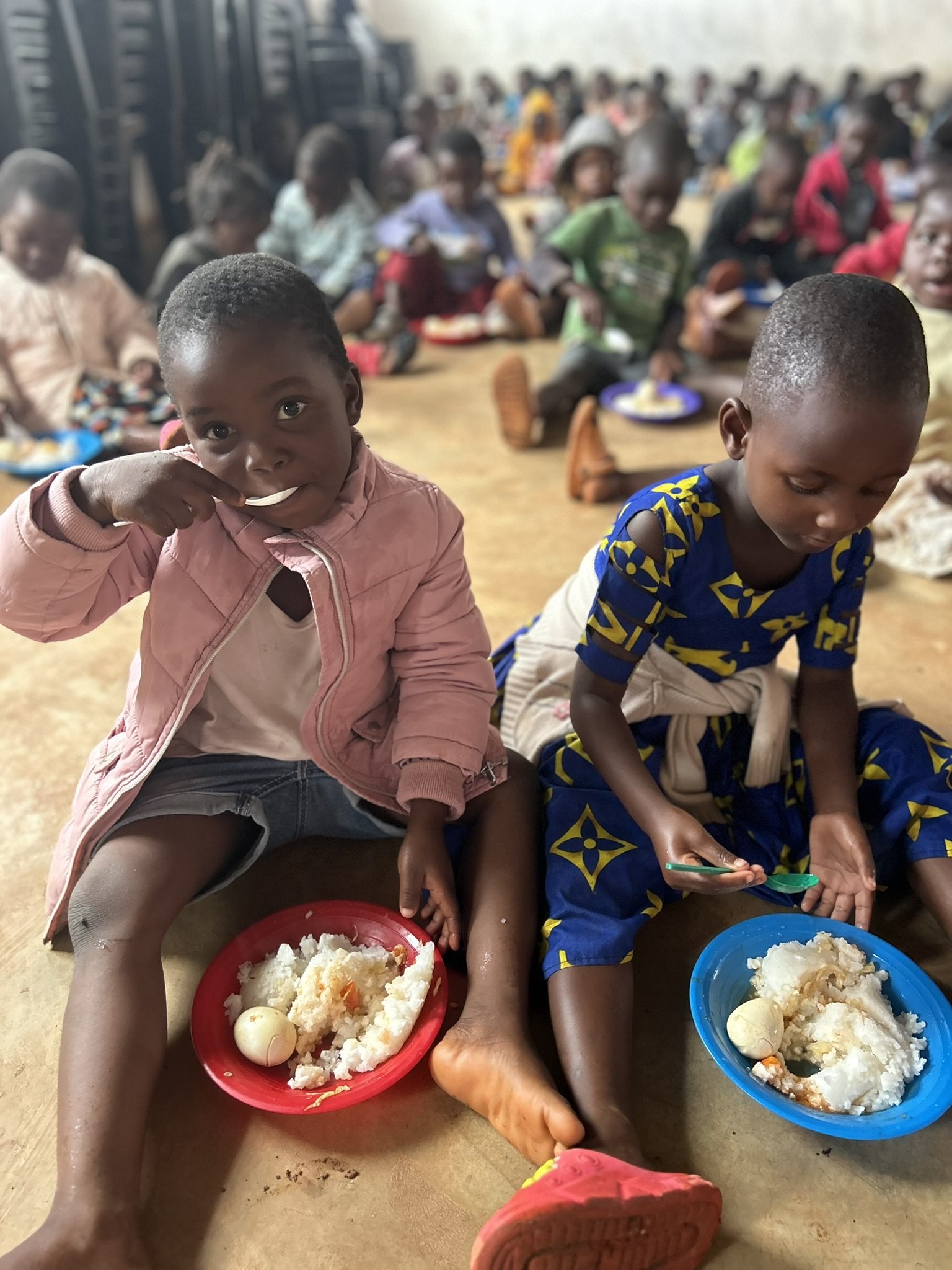 Two young children sitting on the floor eating rice with eggs from colorful plates in a crowded setting with many other children in the background.
