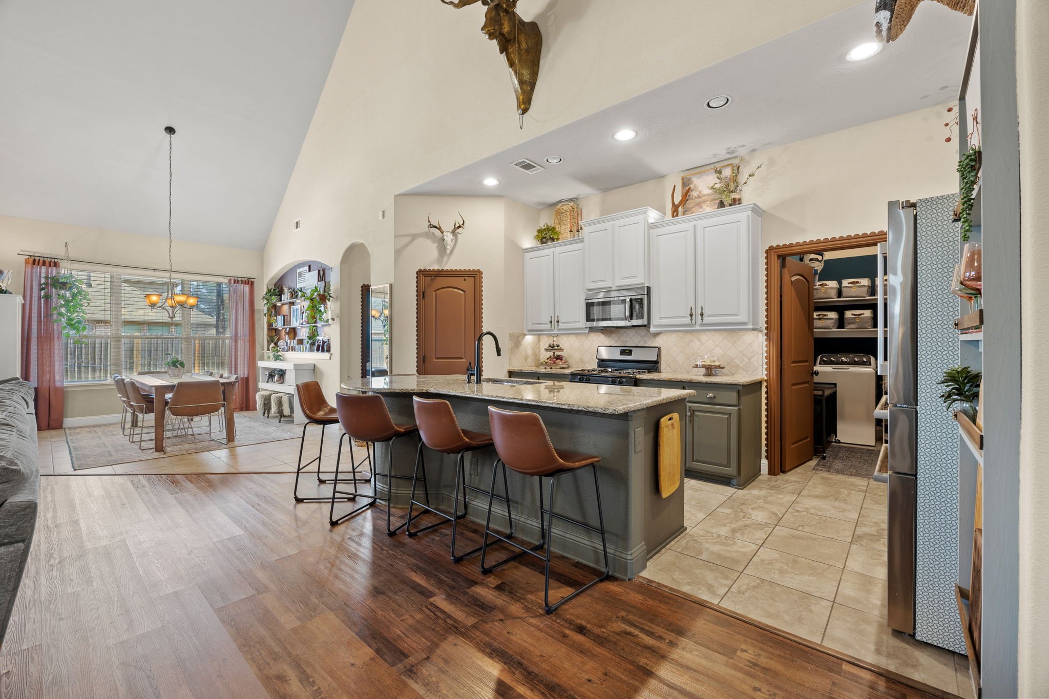 Open kitchen and dining area with vaulted ceiling, white cabinetry, granite countertops, and hardwood flooring. Dining table with six chairs near large window, living room in the background, and kitchen with bar stools and a small pantry.