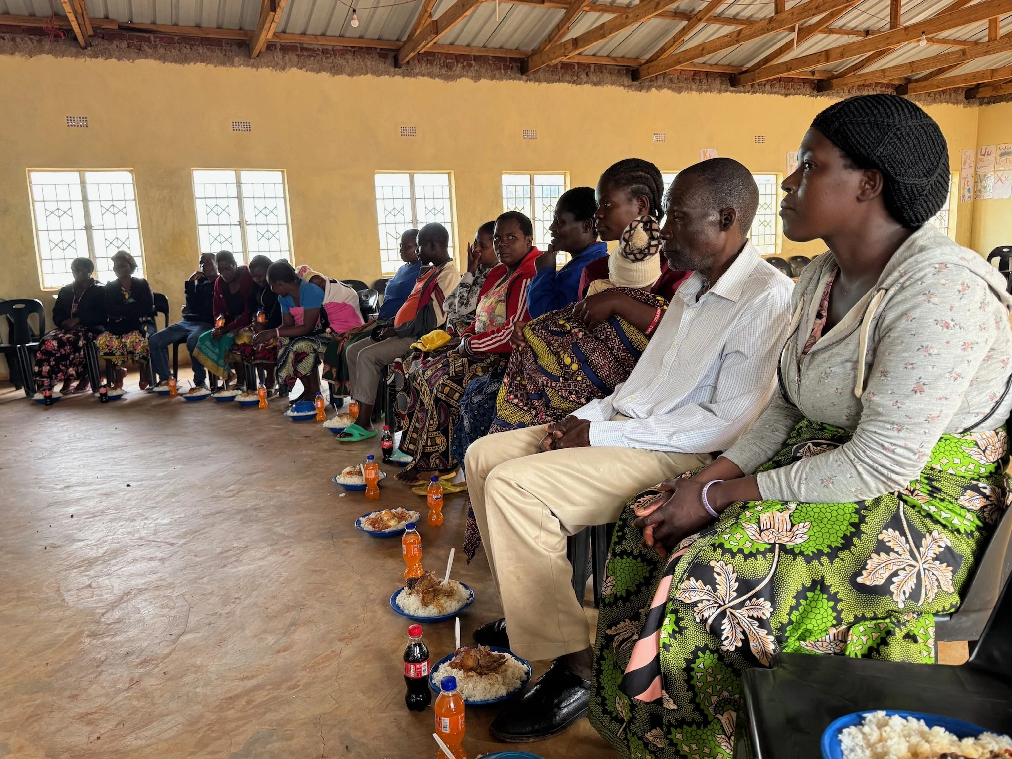 Group of people sitting in a semi-circle inside a room with yellow walls and large windows, having a meal with plates of rice, chicken, and bottles of soda in front of them.