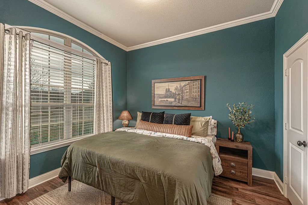 A cozy bedroom with teal walls, a large window with white blinds and patterned curtains, a bed with green bedding and decorative pillows, a wooden nightstand with a lamp and plant, and a framed artwork above the bed.
