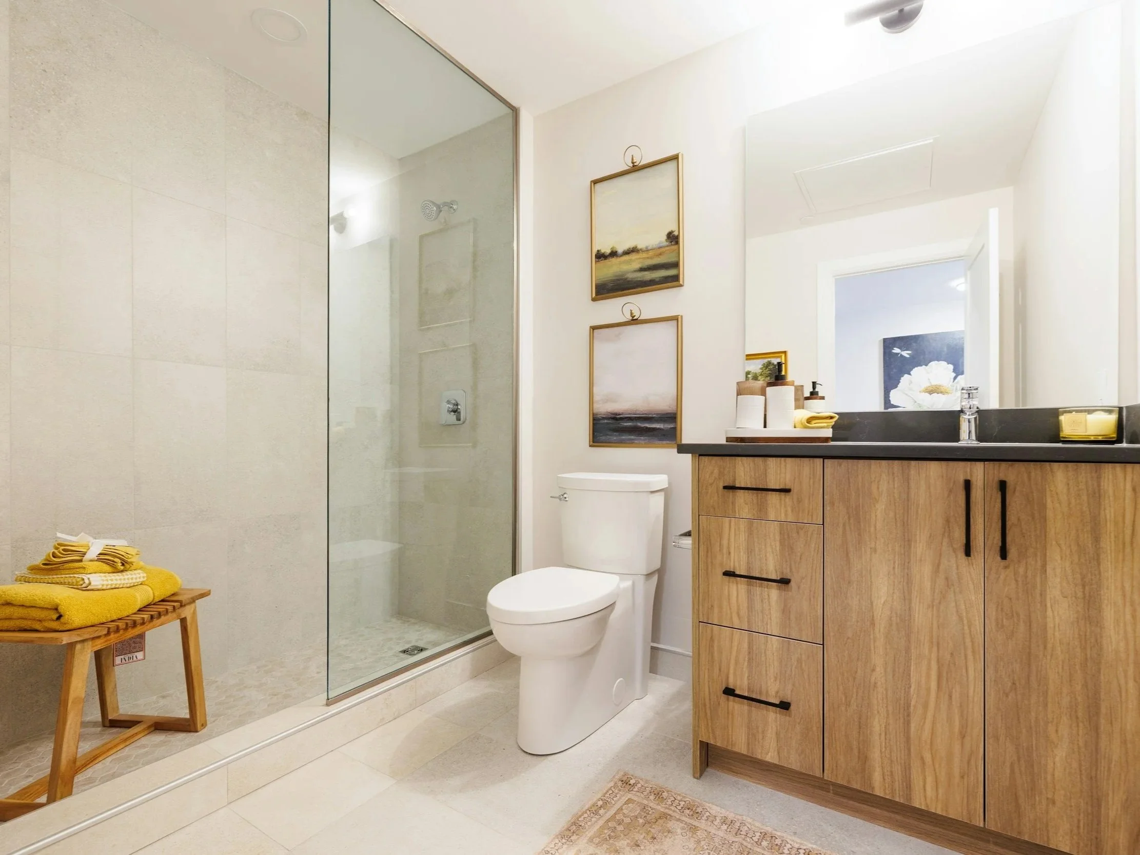 Bathroom with a glass shower enclosure, white toilet, wooden vanity with black countertop, large mirror, and framed artwork on the wall.
