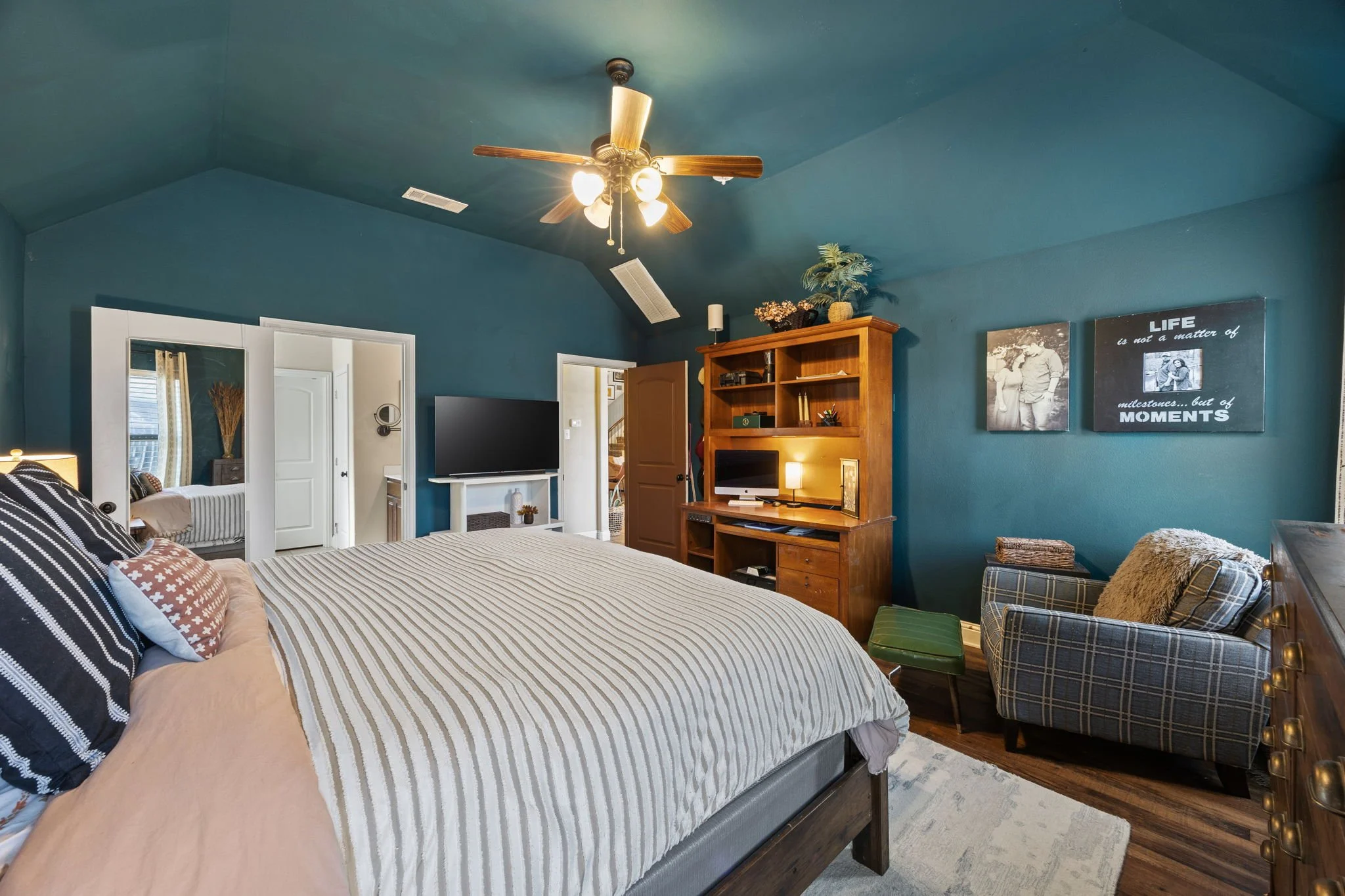 A bedroom with teal walls, a large bed with striped bedding, a TV on a stand, a wooden bookshelf with a computer, a plaid armchair with a pillow, and wall art that reads 'Life is not a matter of moments'.