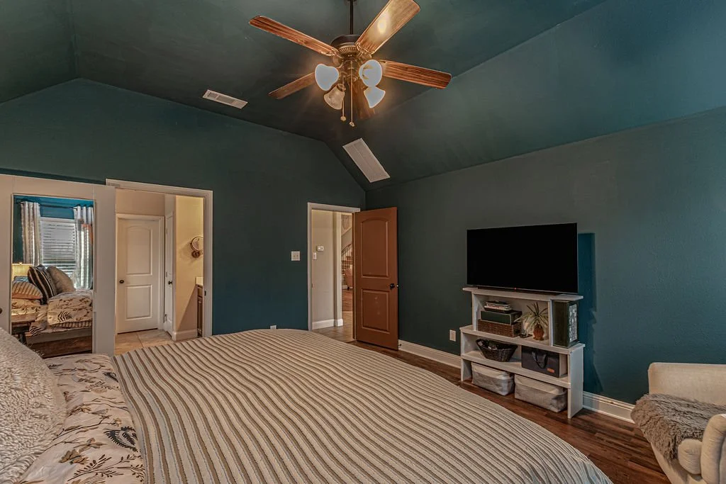 A bedroom with teal walls, a bed with striped bedding, and a ceiling fan with wooden blades and lights. There's a television on a white stand, an open door leading to another room, and a glimpse of an adjoining room with a bed and window.