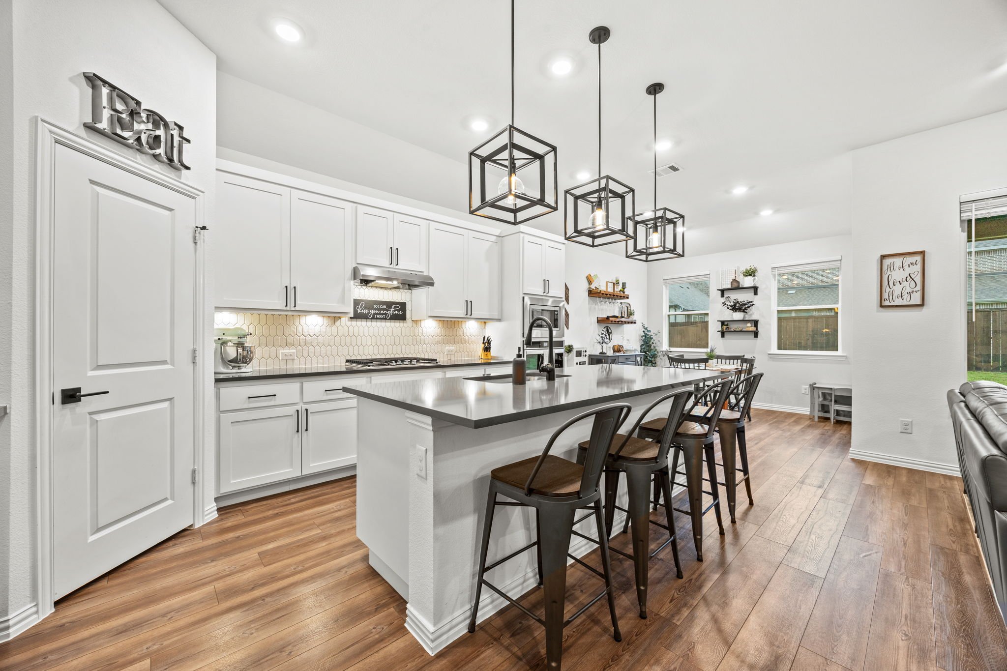 Modern kitchen with white cabinets, a large island with a gray countertop, black metal bar stools, pendant lighting, a honeycomb tile backsplash, and wooden flooring.