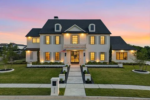 Large two-story house with a manicured lawn and a sidewalk leading to the front door, illuminated windows, and a sunset sky in the background.