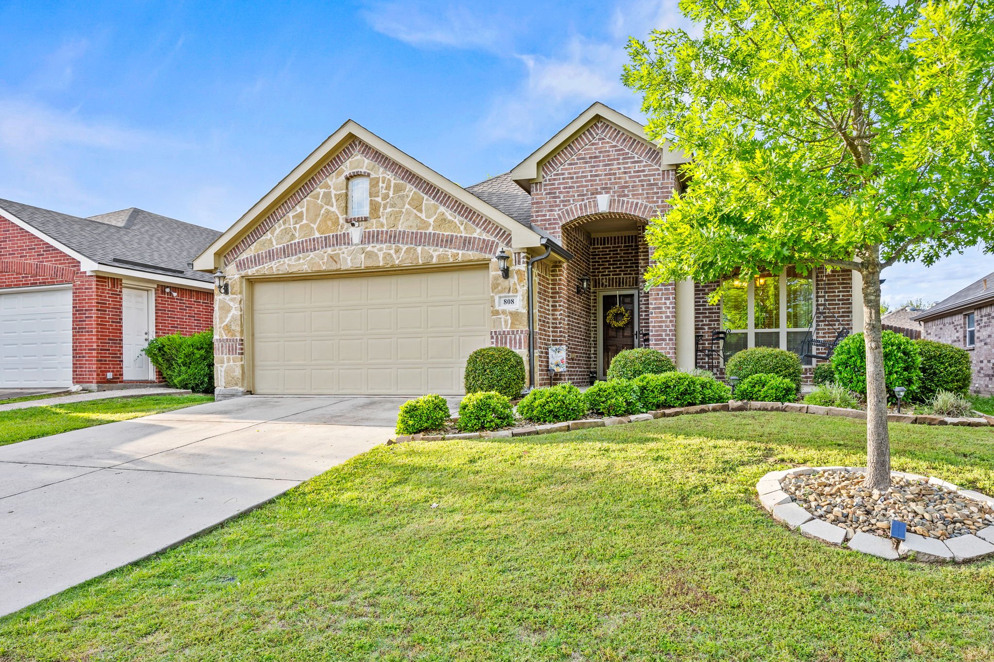A suburban house with a brick and stone exterior, a front lawn with neatly trimmed bushes, and a driveway leading to a garage.