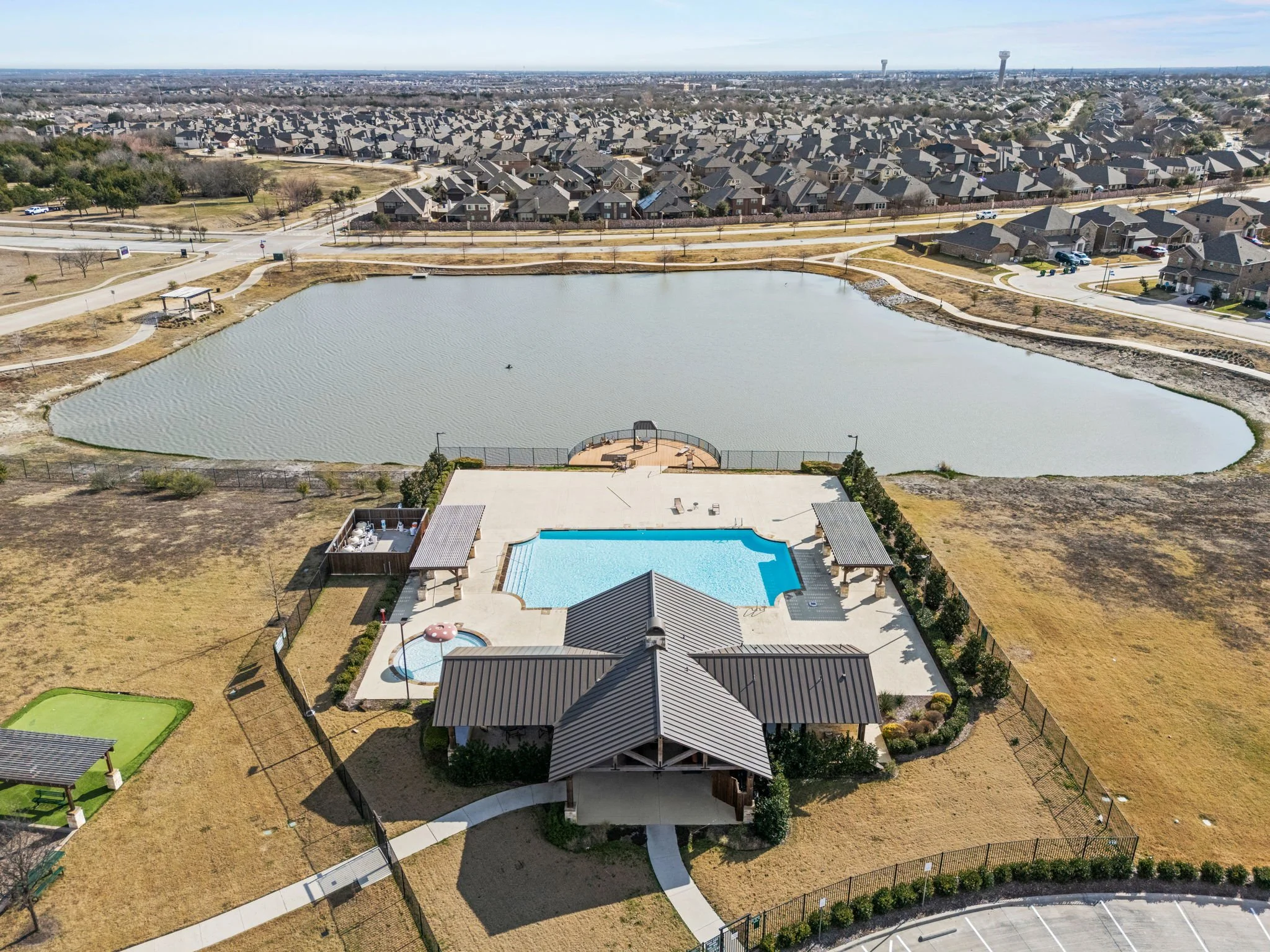 Aerial view of a community swimming pool, a smaller hot tub, a pond with a walking trail, and a residential neighborhood in the background.
