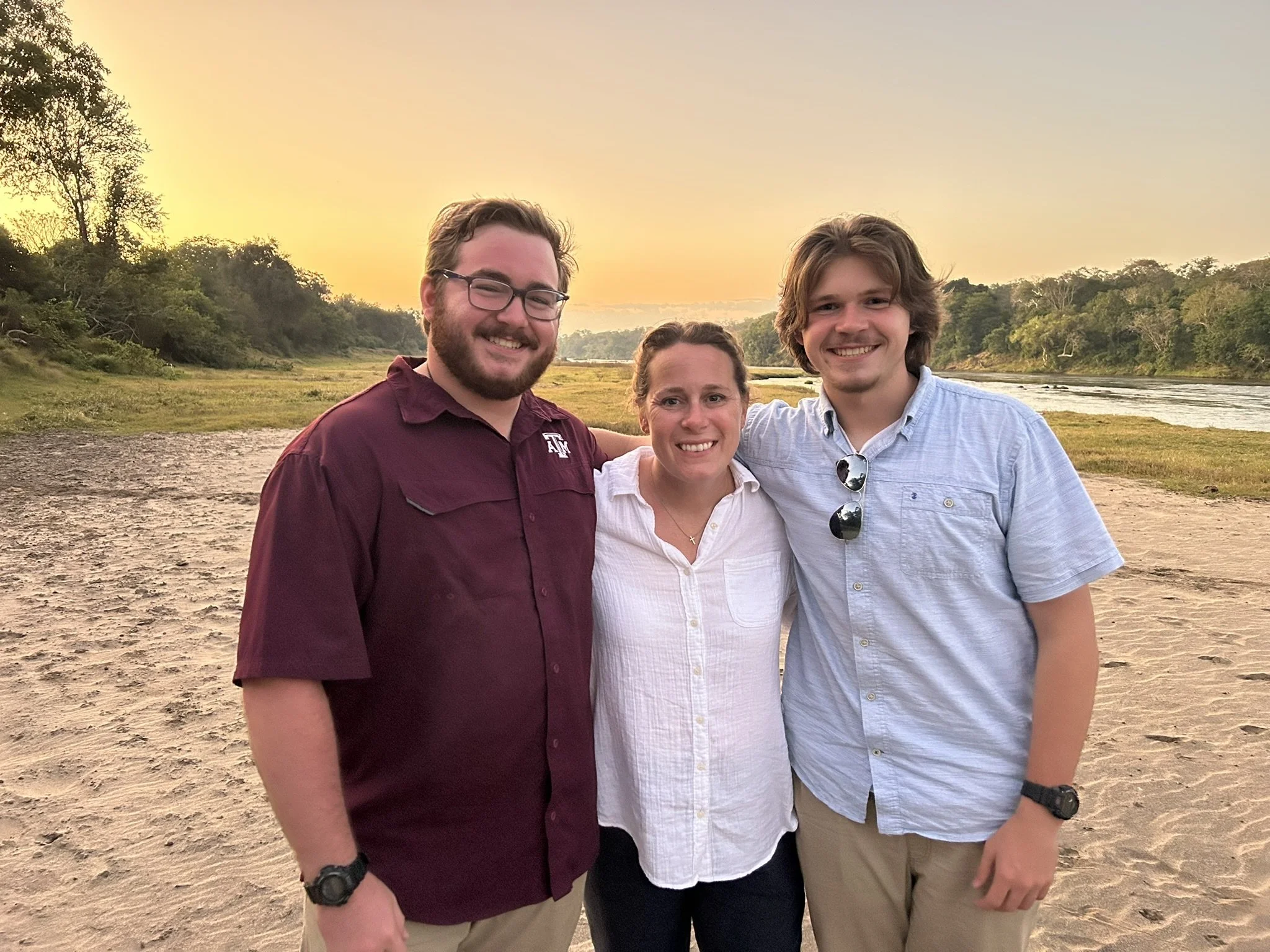 Three people smiling and standing close together on a sandy riverbank during a sunset, with trees and a river in the background.