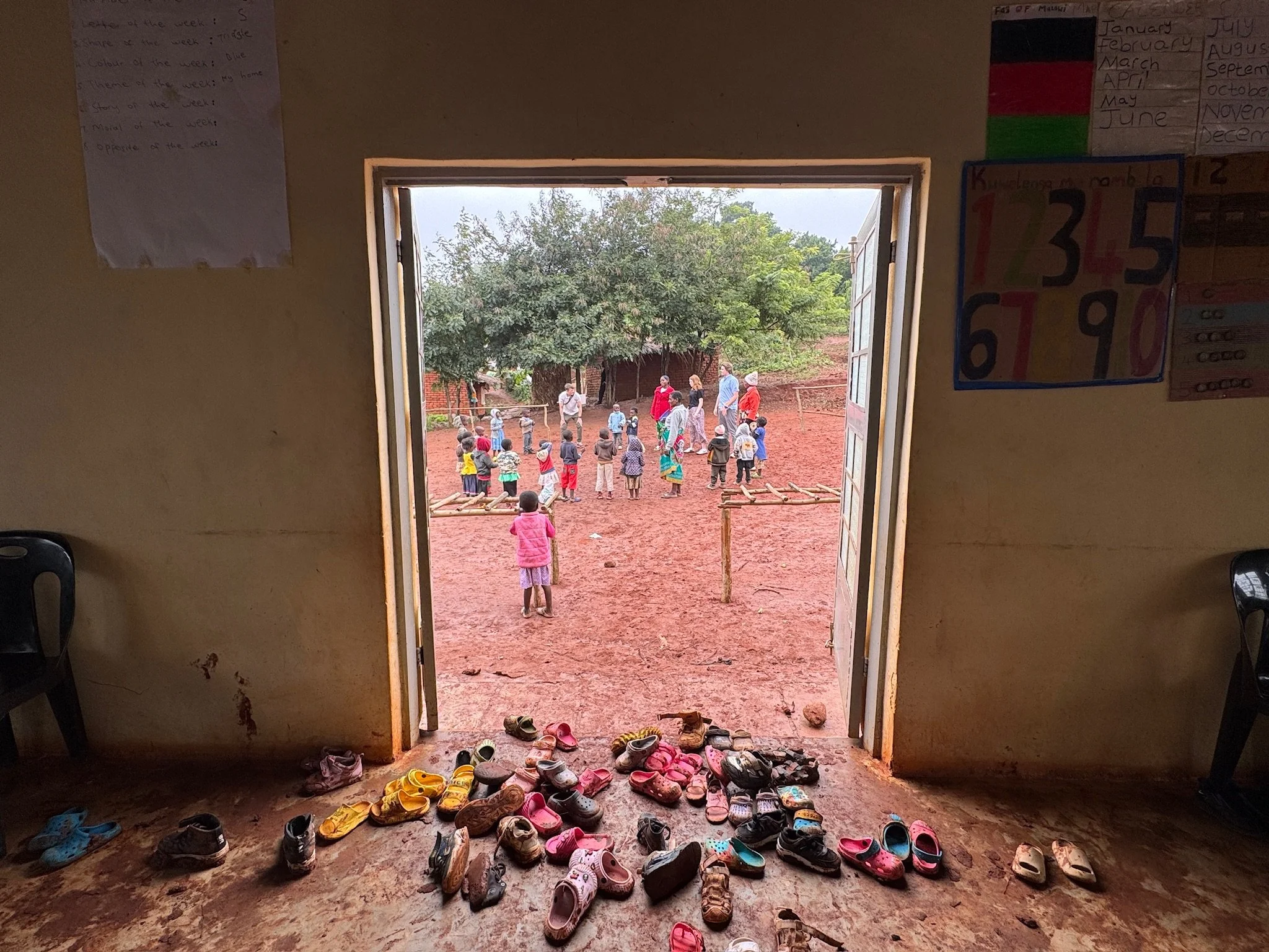 View through an open door showing children and adults outside on a red dirt ground, with trees in the background. Inside, there are many pairs of children's shoes on the floor near the door, and educational posters on the walls.