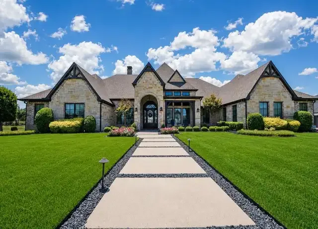 Large stone house with a landscaped front yard and a stone pathway leading to the front door, under a blue sky with clouds.