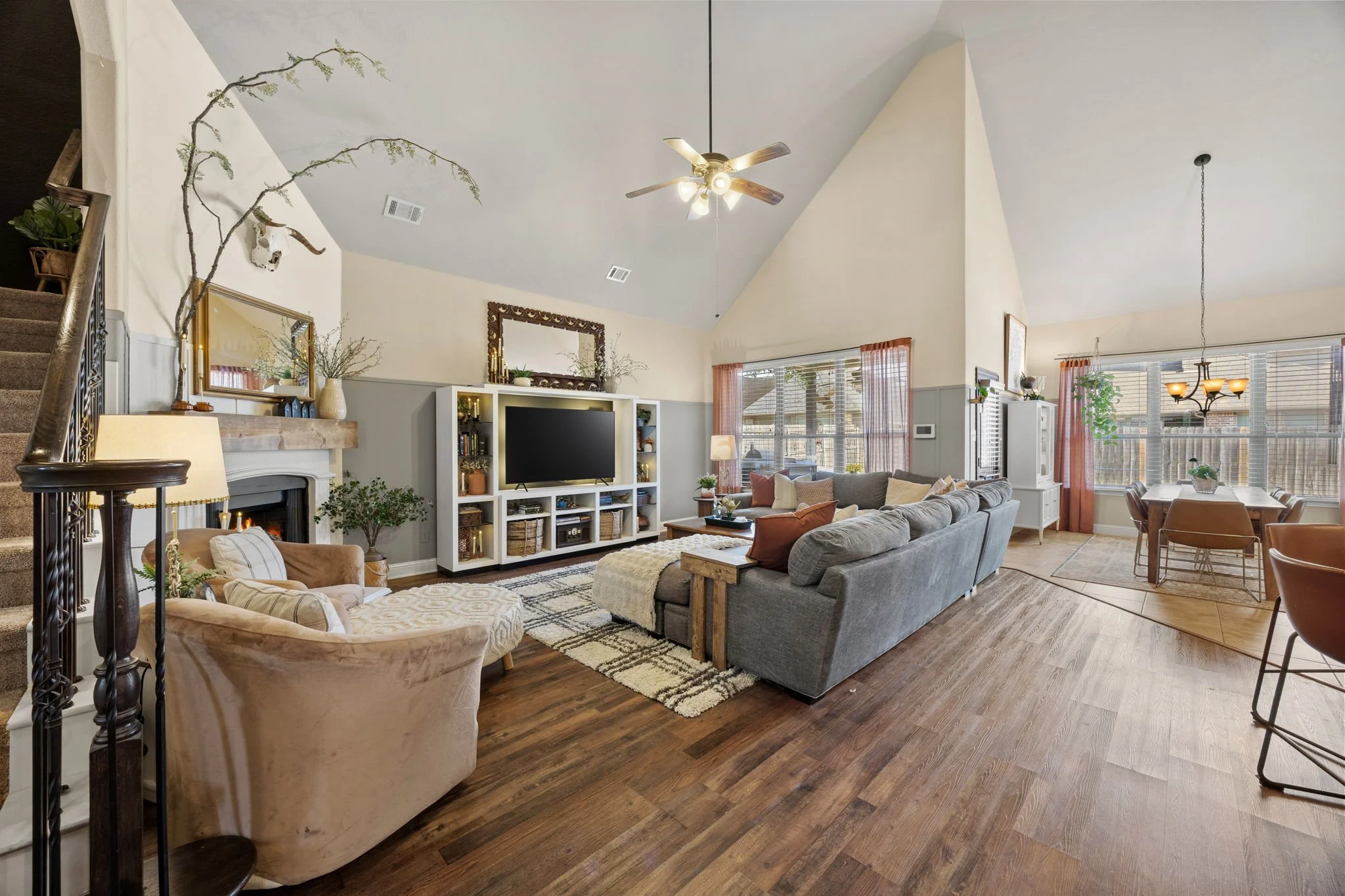 Spacious living room with a vaulted ceiling, large gray sofa, beige armchair, and a white entertainment center with a TV, surrounded by windows and a dining area in the background.