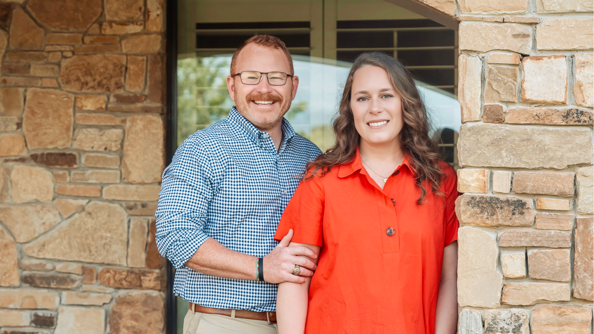 a man with glasses and a woman with curly brown hair standing outside in front of a stone wall, smiling at the camera