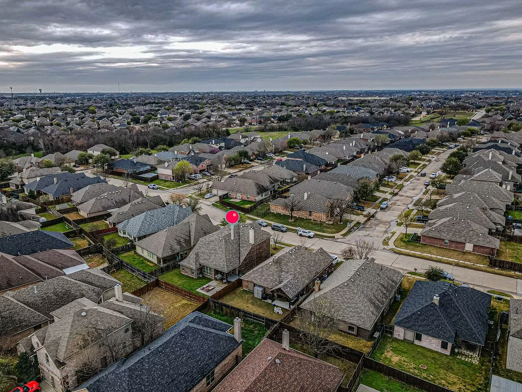 Aerial view of a suburban neighborhood with rows of houses, streets, and cars, under a cloudy sky.
