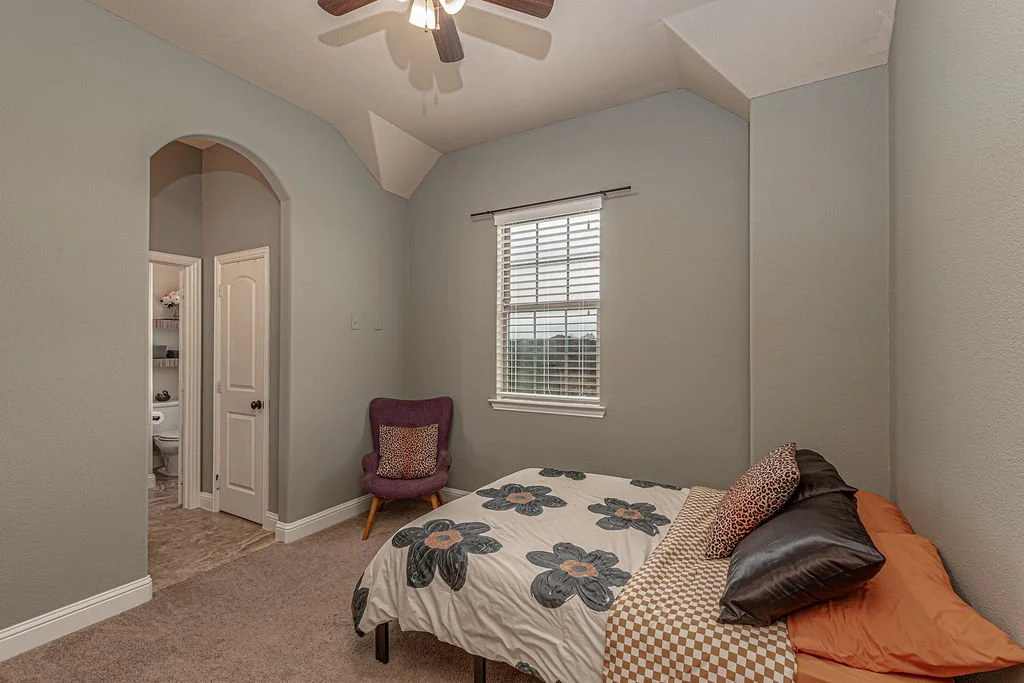 A bedroom with a bed, purple armchair, window with blinds, ceiling fan, and an open door showing a bathroom.