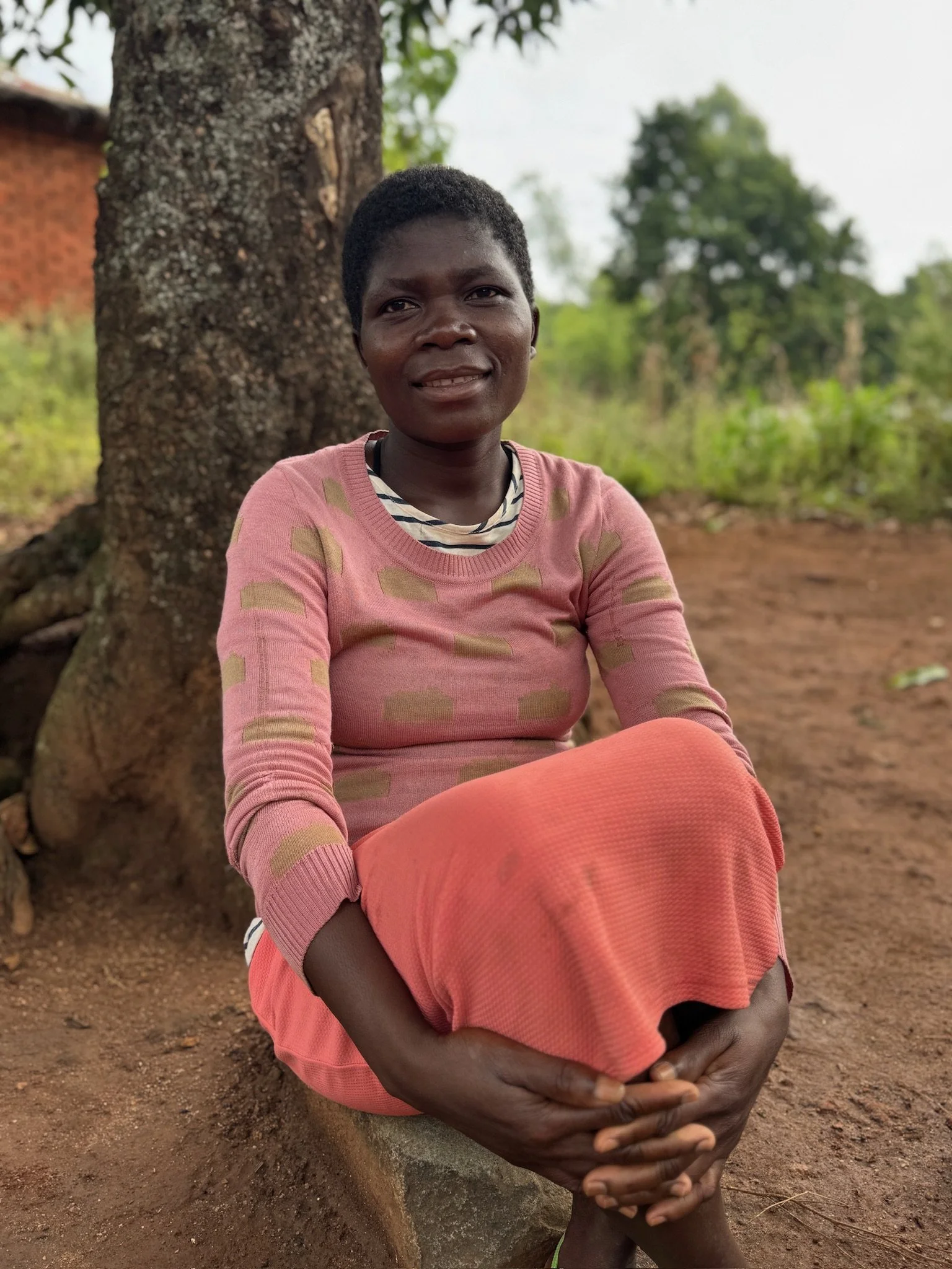 A woman sitting outdoors on a dirt ground, leaning against a tree, with a background of greenery and a brick building, wearing a pink patterned long-sleeve shirt and a coral skirt.