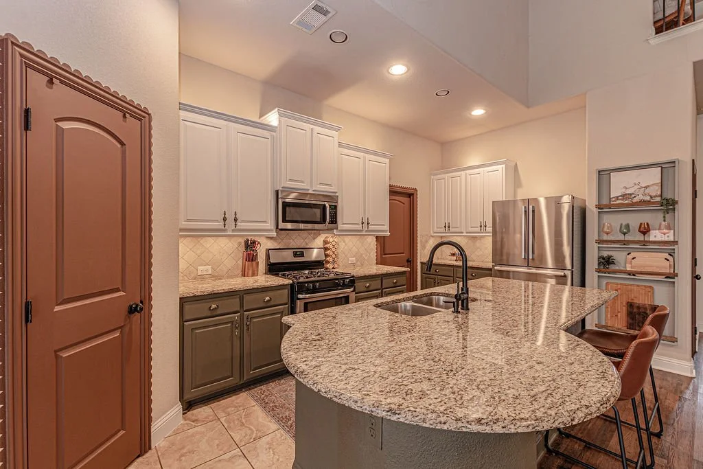 A modern kitchen with granite countertops, white and olive green cabinets, stainless steel appliances, and a small bar area with two chairs.
