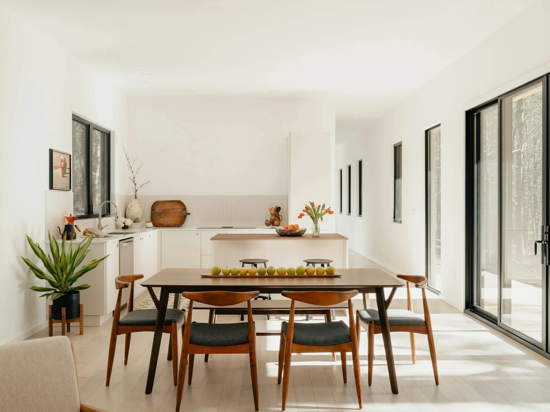 Bright, minimalist kitchen and dining area with white walls, large windows, wooden dining table with six chairs, and decorative plants and artwork.