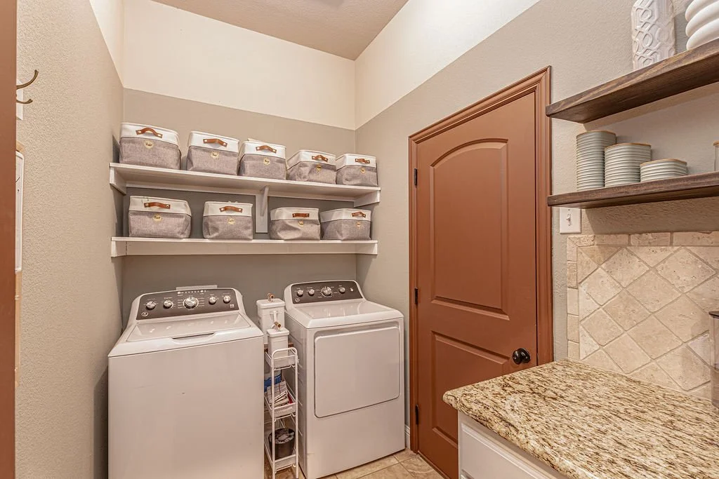 Laundry room with top-loading washing machine and dryer, shelves with laundry baskets, a granite countertop, and open shelves with cups and bowls.