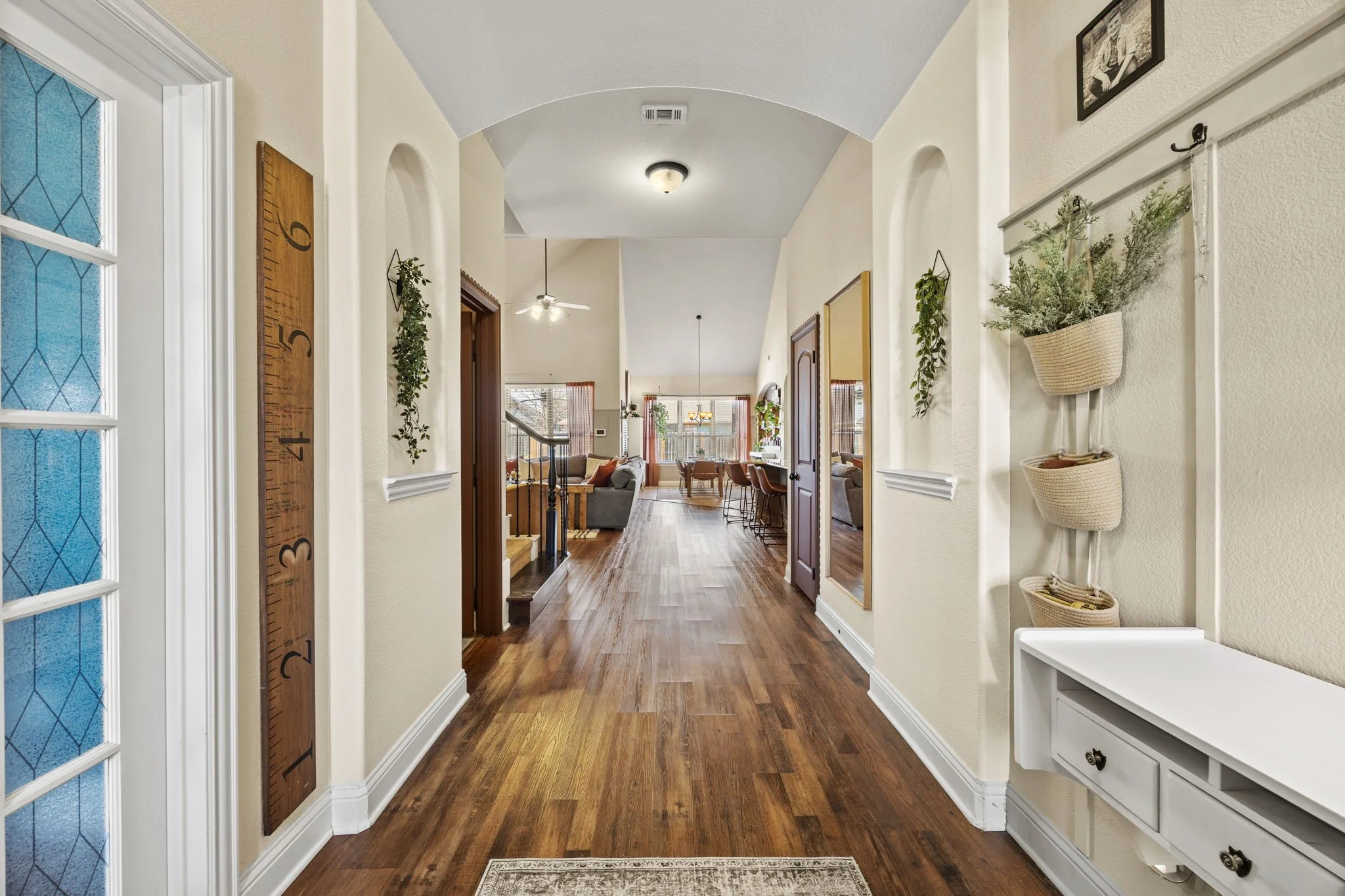 View of a hallway in a house with hardwood floors, decorated with hanging plants, a large wooden growth chart on the wall, and framed pictures. The hallway opens into a living and dining area with large windows, a ceiling fan, and various furniture.