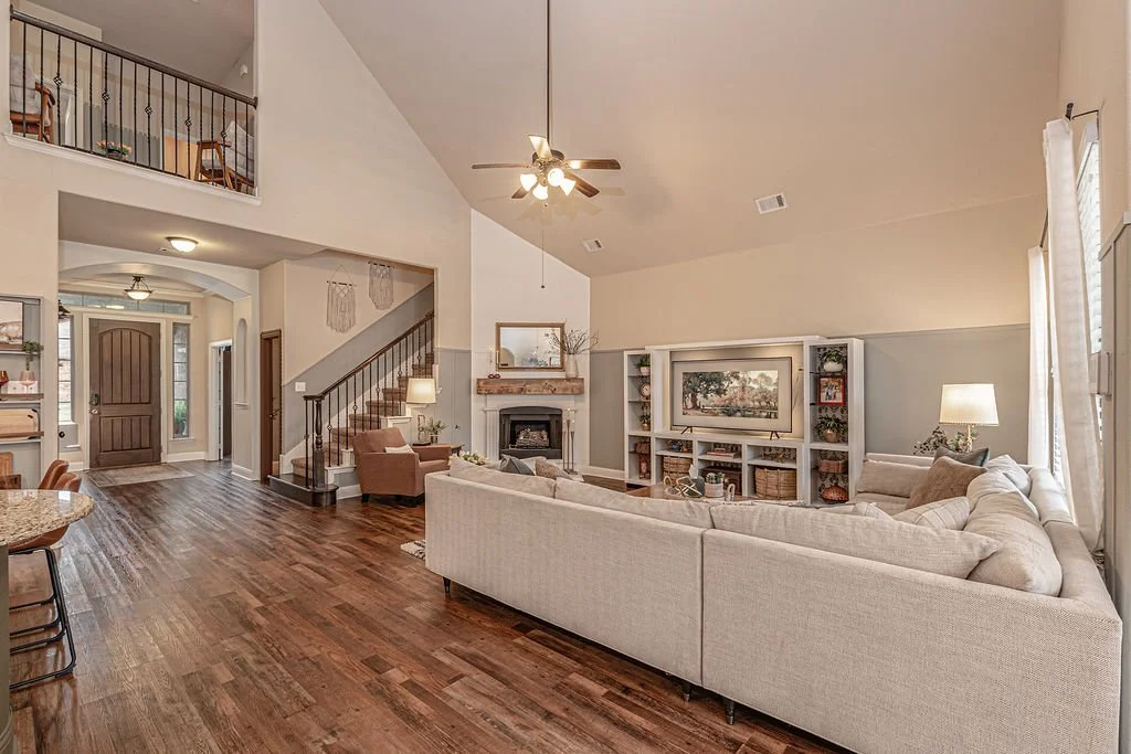 Living room with beige sectional sofa, TV on white entertainment center, fireplace, wooden flooring, and large windows with white curtains.