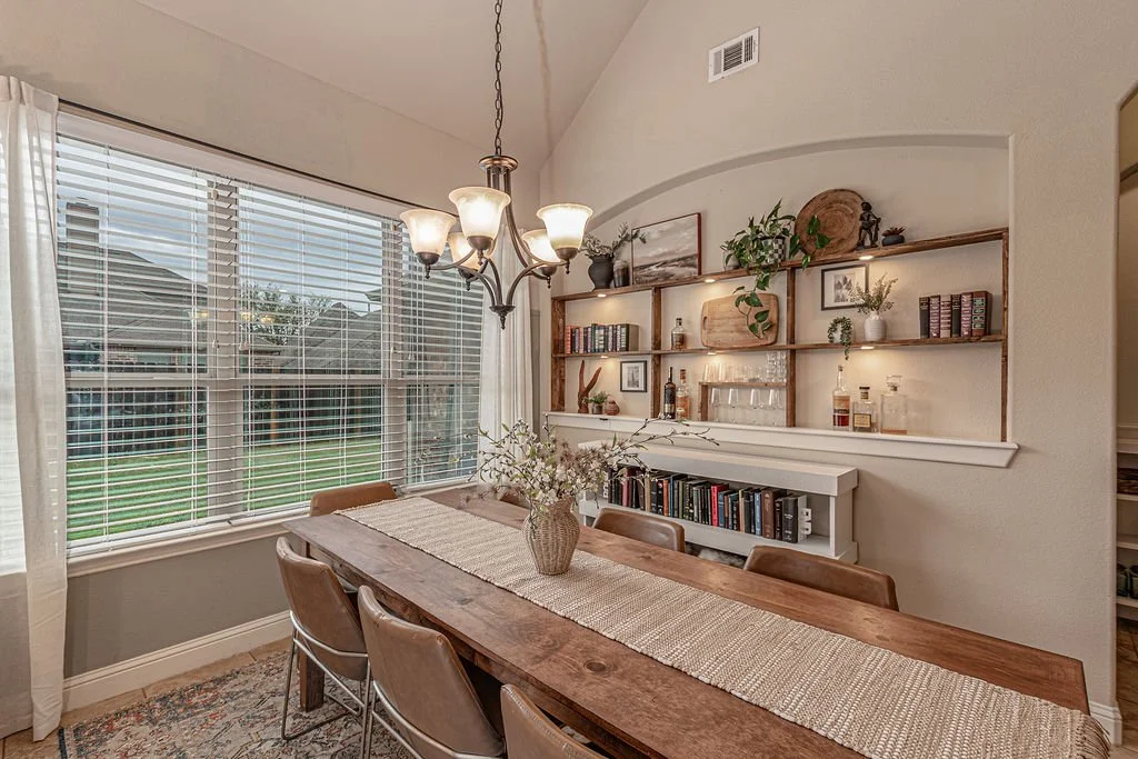 A dining room with a wooden table, six chairs, a vase with flowers, large windows with blinds, a chandelier, and built-in wooden shelves with books, decorative items, and plants.