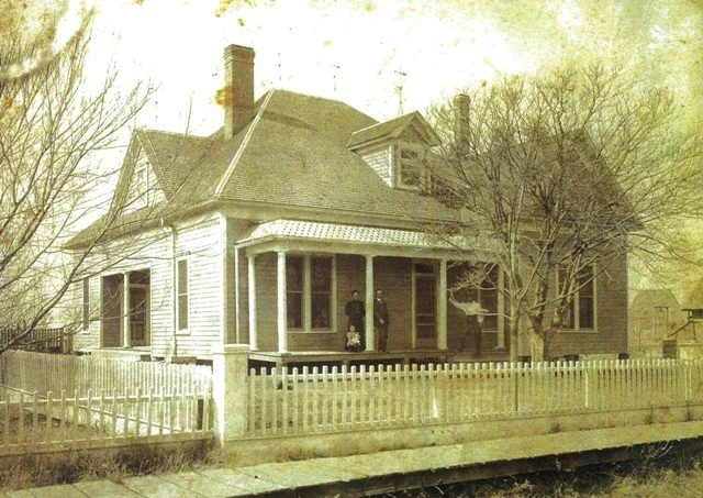 A vintage-style photograph of a large, old-fashioned house with a wraparound porch, white picket fence, and leafless trees in front. There are two people standing on the porch.