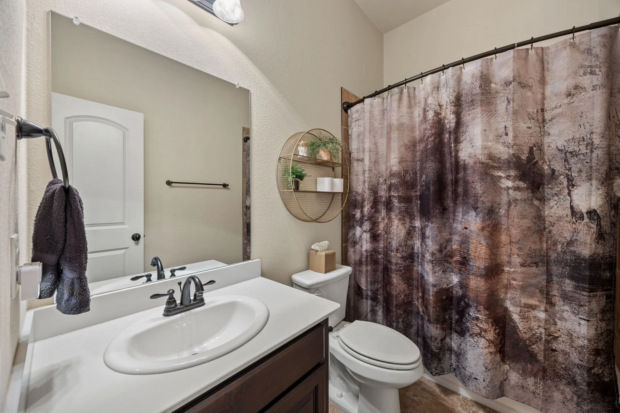 A small bathroom with a white vanity, a mirror, a toilet, a wall-mounted shelf with plants and tissue paper, a brown and black shower curtain, and a beige wall.