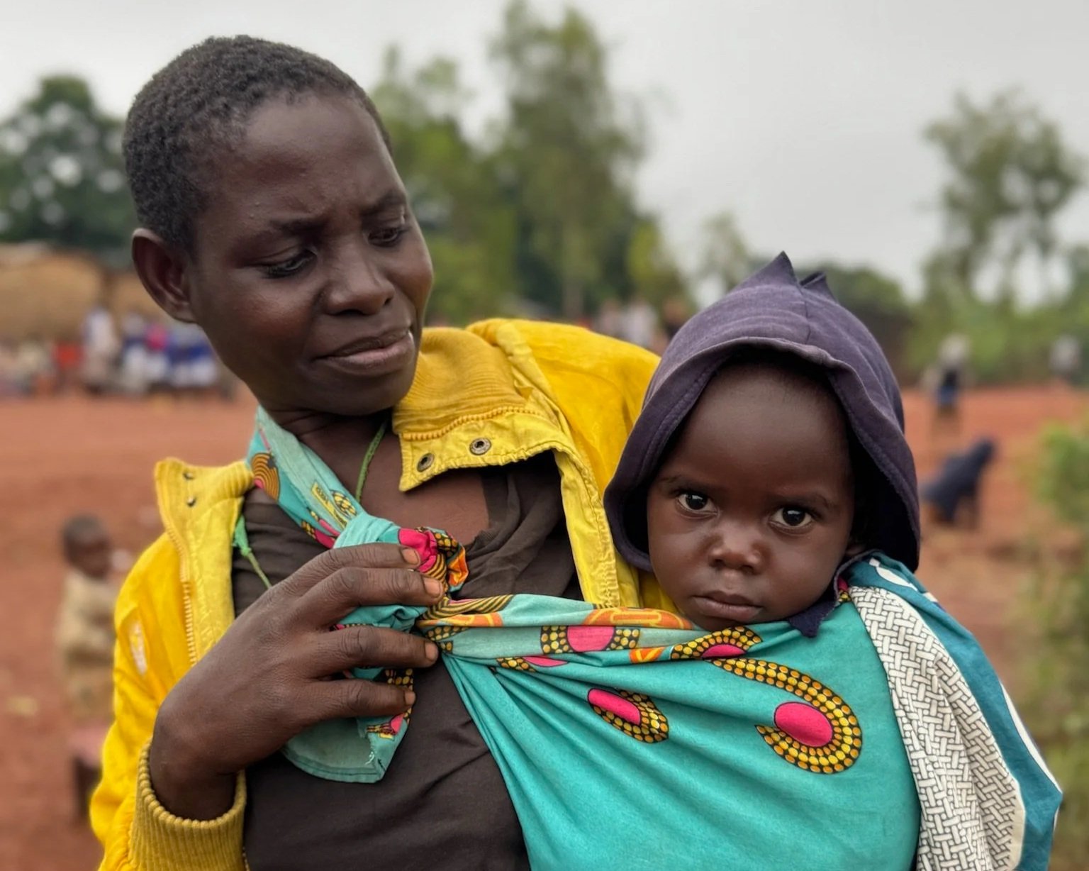 A woman holding a young child in a colorful wrap, outdoors in a rural area with trees and other people in the background.