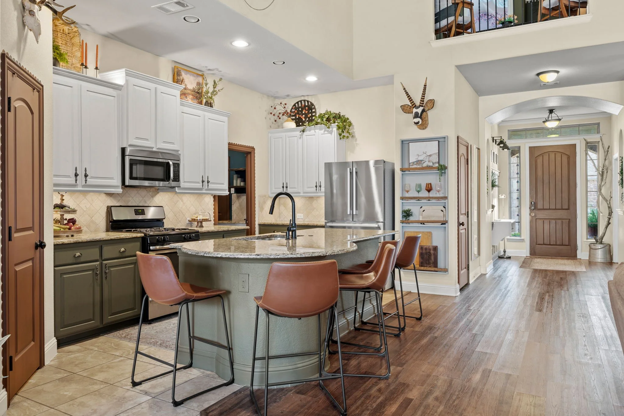 A spacious kitchen with a central island, white and green cabinets, stainless steel appliances, and hardwood floors leading to an entryway with a front door. Decor includes plants, art, and a mounted animal head.