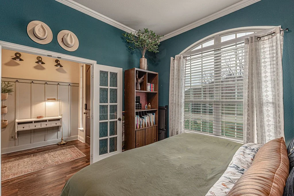 A bedroom with a large window, bed, bookshelf, and decorative hats on the wall.