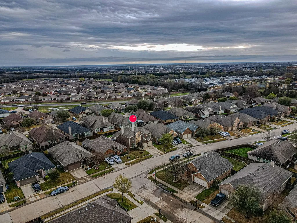 Aerial view of a suburban neighborhood with houses, streets, and cars, under a cloudy sky.