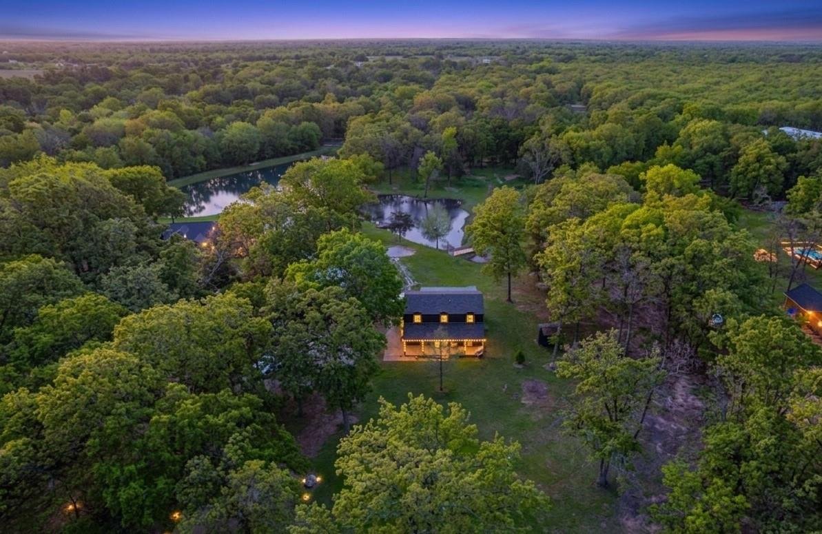 Aerial view of a lush green park with ponds and trees, a house with lit windows in the center, and other houses nearby, at sunset.