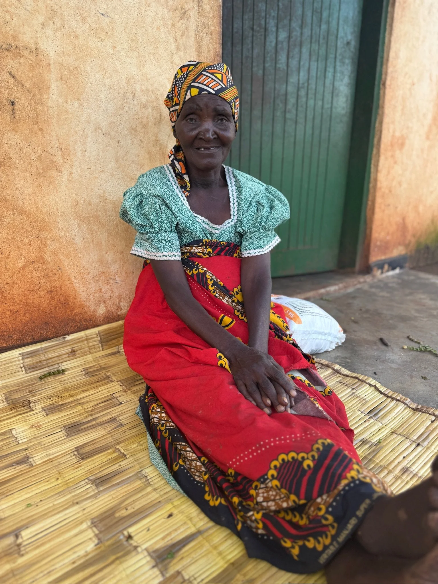 An elderly woman sitting on a woven mat outside, wearing colorful traditional clothing and a patterned headscarf, smiling at the camera.
