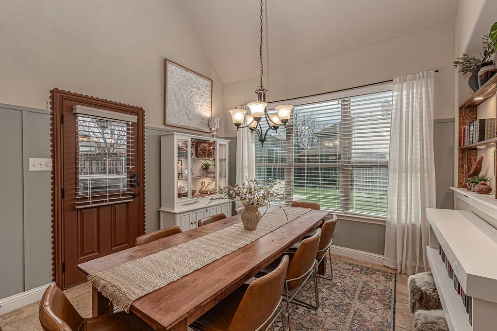 Dining room with wooden table and brown chairs, a chandelier, a white cabinet, large window with white curtains, and a bookshelf.