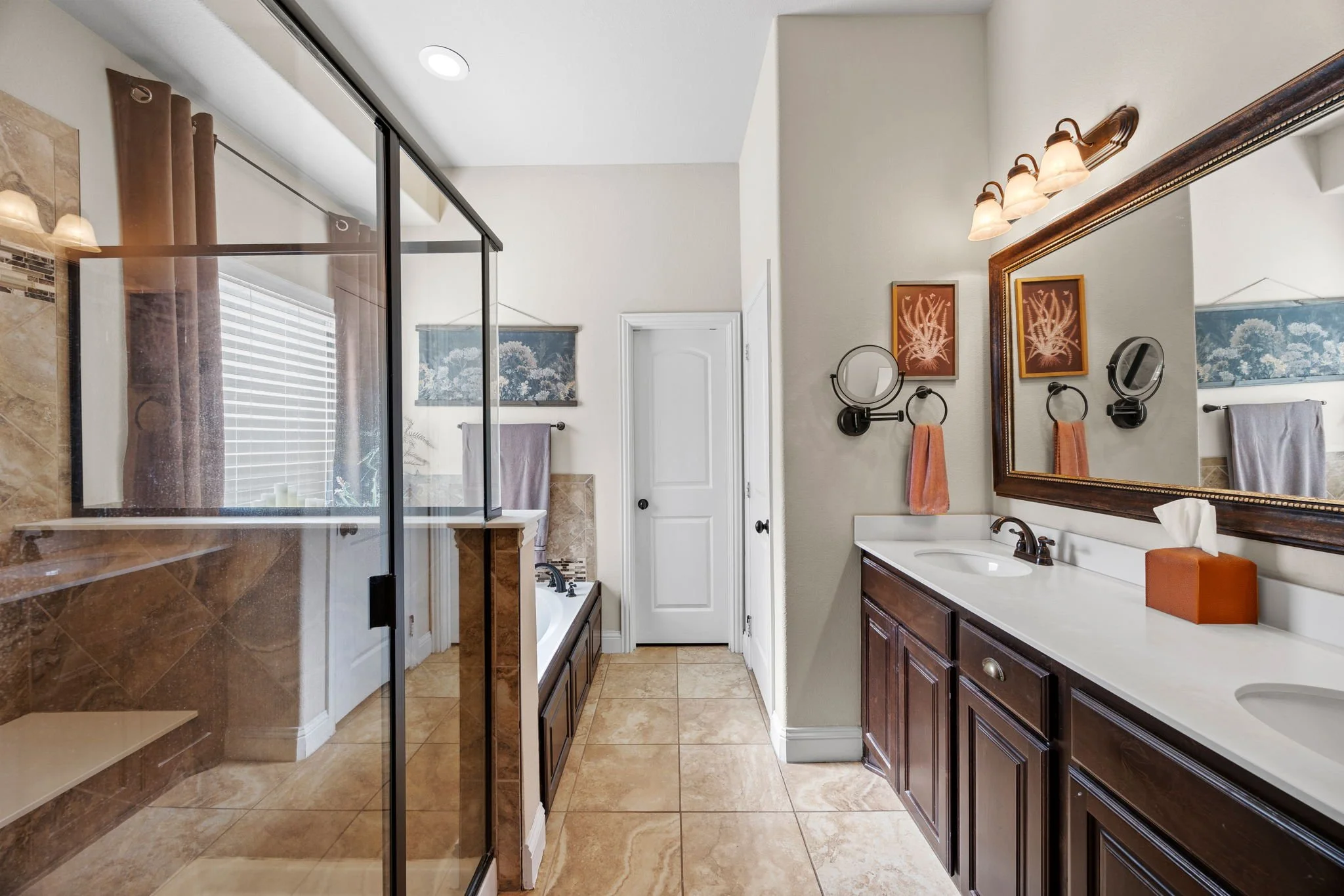 A spacious bathroom with a glass-enclosed shower, a large mirror, two sinks on a dark wood vanity, a bathtub with a window and curtains, and wall decorations, with tan tiled floors and cream-colored walls.
