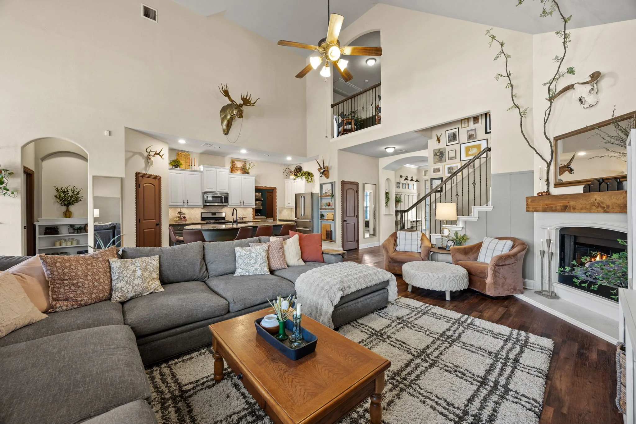 Spacious living room with vaulted ceiling, gray sectional sofa, and wood coffee table. Fireplace with a wooden mantle, wall decor, and climbing plant. Open kitchen with white cabinets, stainless steel appliances, and bar seating. Armchairs, area rug,