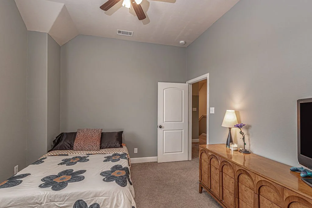 A bedroom with a bed, a dresser with a lamp, a small TV, and a doorway, with walls painted in light gray and a ceiling fan.