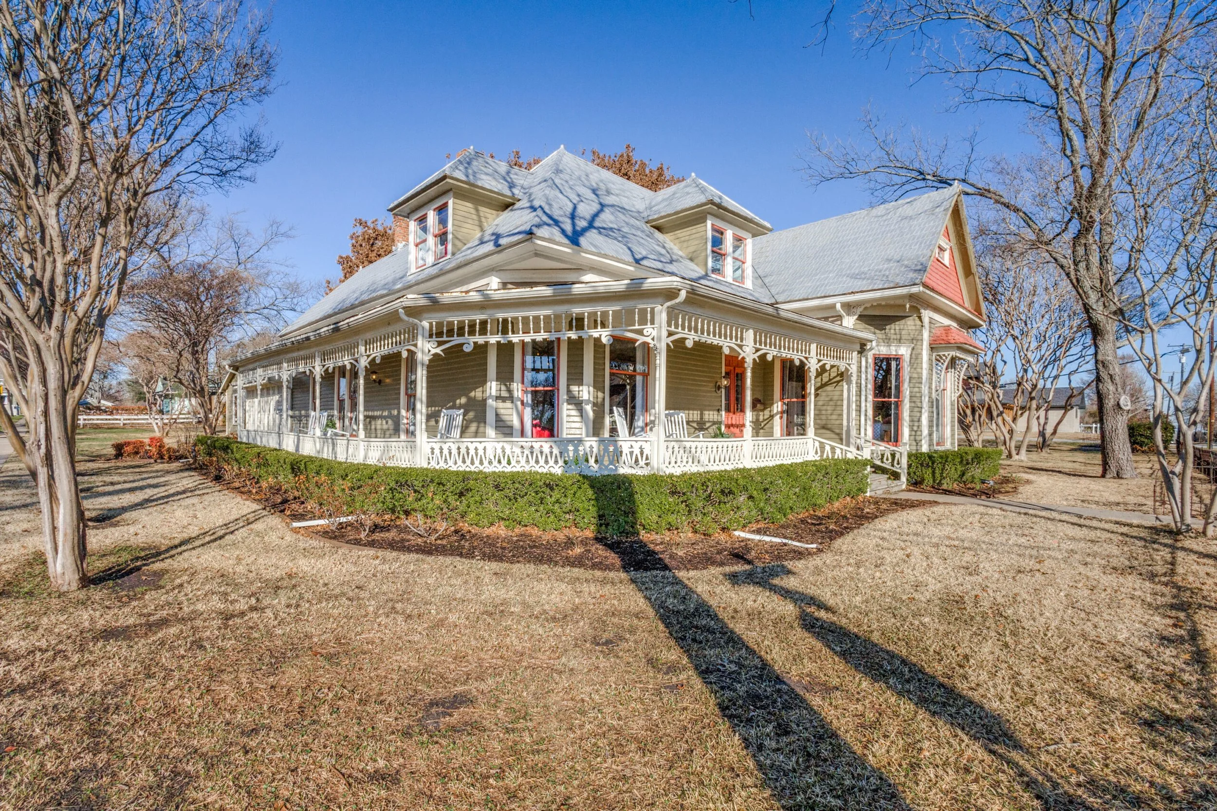 A large vintage Victorian house with a wraparound porch, light green exterior, ornate trim, and a gabled roof with dormer windows, surrounded by a yard with leafless trees and a clear blue sky.