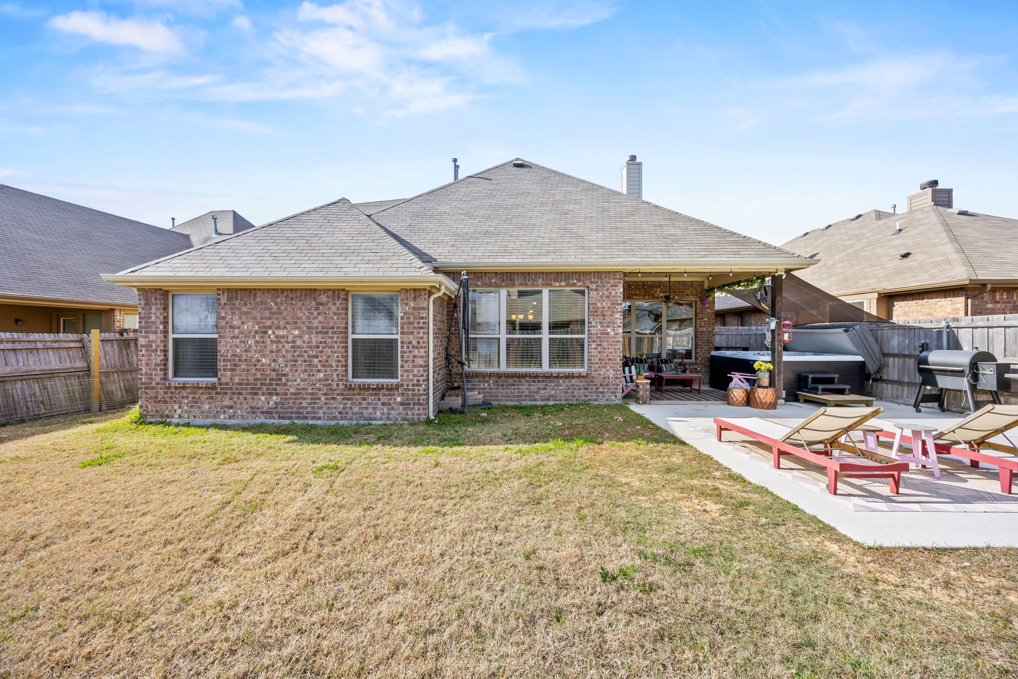 Backyard patio area with lounge chairs, a grill, and seating area under a covered porch, with a fence and neighboring houses in the background.