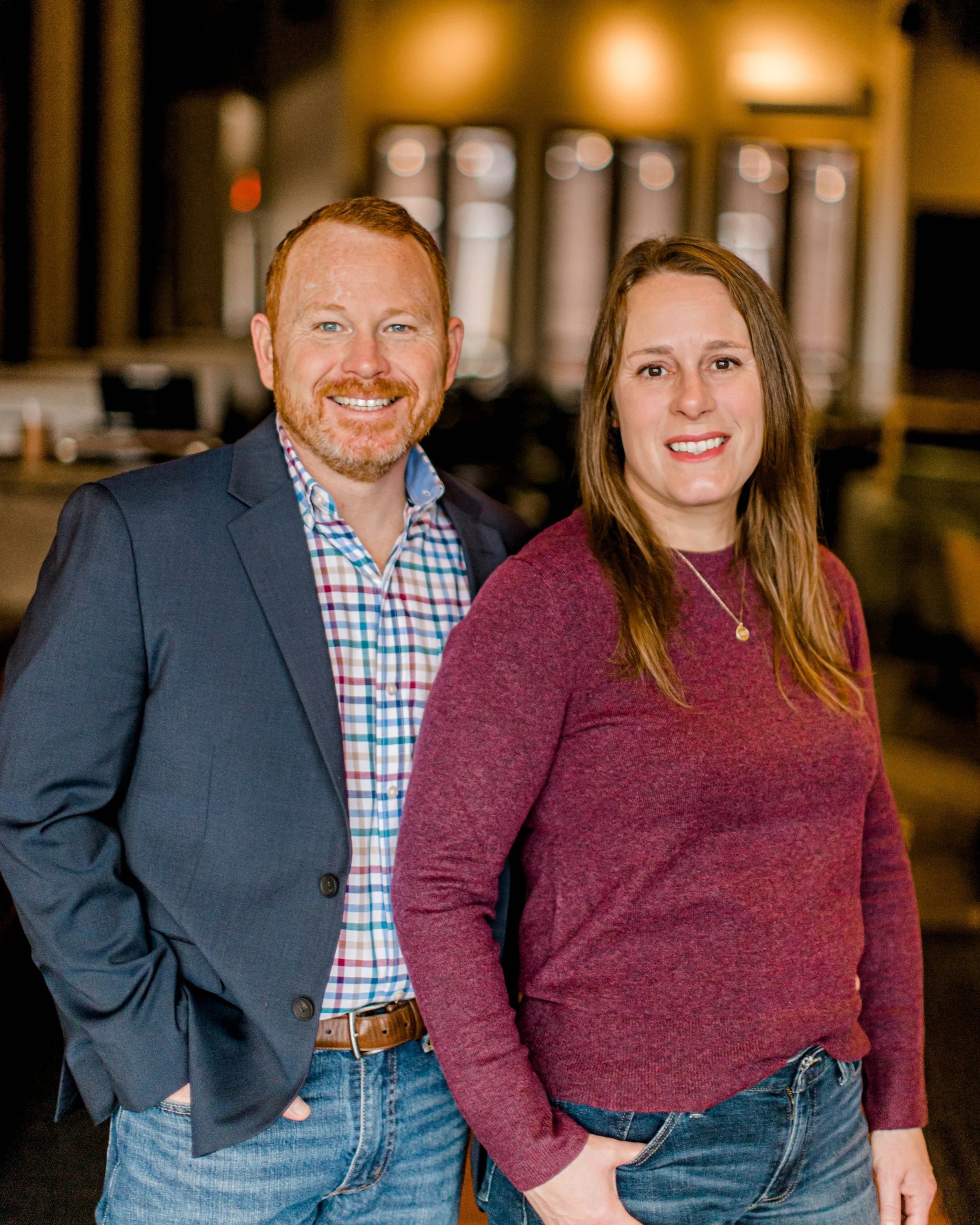 A man in a blue blazer and checkered shirt standing next to a woman in a maroon sweater inside a warmly lit room with wooden decor and blurred background.