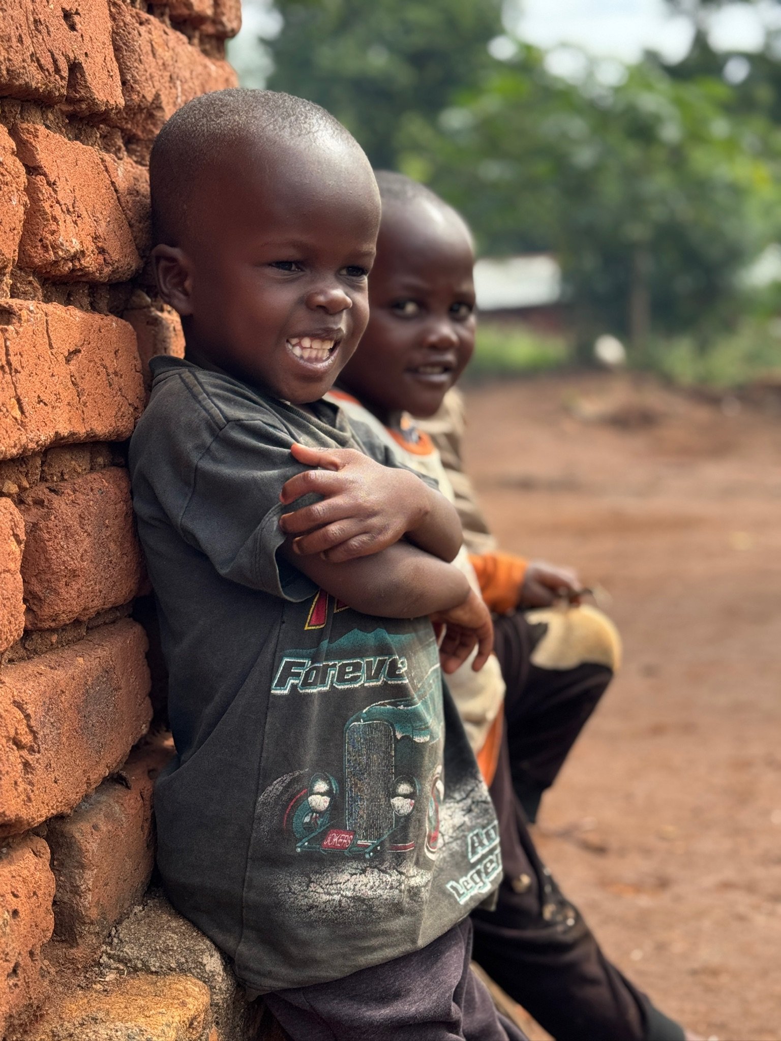 Two young boys sitting outdoors, leaning against a brick wall, with the boy in the foreground smiling and crossing his arms. The other boy is sitting behind him, looking at the camera. The setting appears to be rural with greenery in the background.