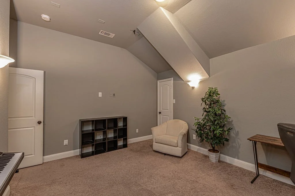 A cozy room with gray walls, a beige armchair, a tall potted plant, a small wooden desk, a black storage cube, a doorway, and a keyboard partially visible on the left. The ceiling is sloped with a textured finish, and warm lighting illuminates the sp