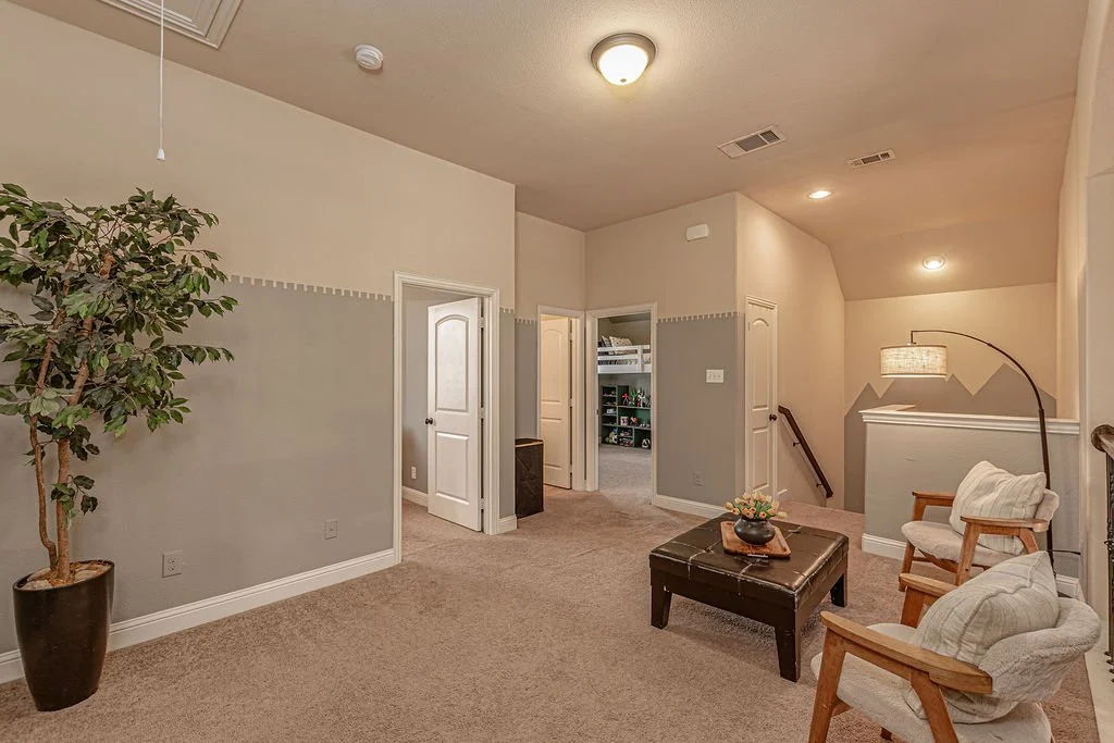 Living room with beige carpet, white walls, a potted plant, a black ottoman, two wooden chairs with cushions, a floor lamp, and a staircase with a wooden handrail.