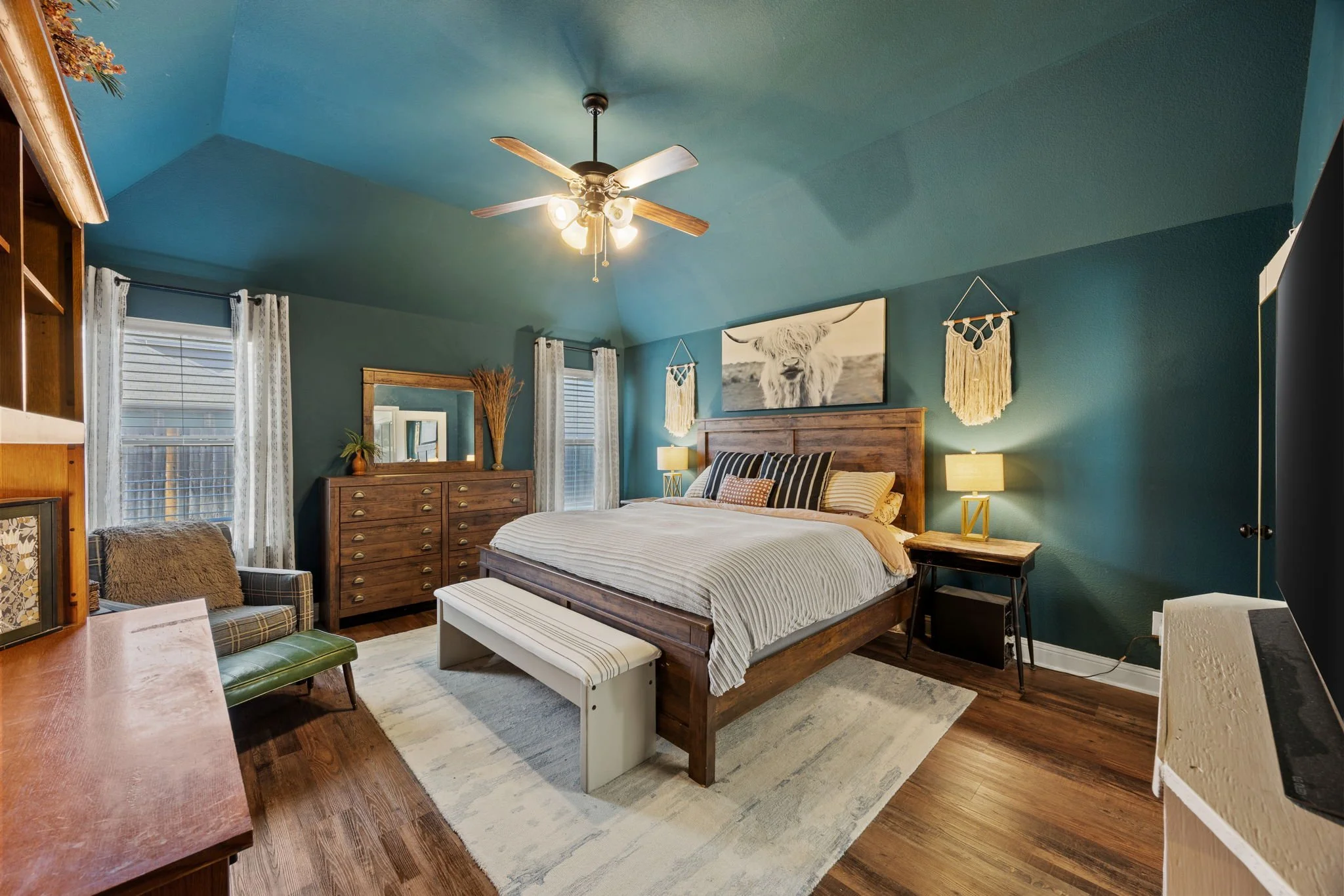 Bedroom with a wooden bed, blue walls, a ceiling fan, two windows with white curtains, a chest of drawers, a mirror, an armchair, and decorative wall hangings.