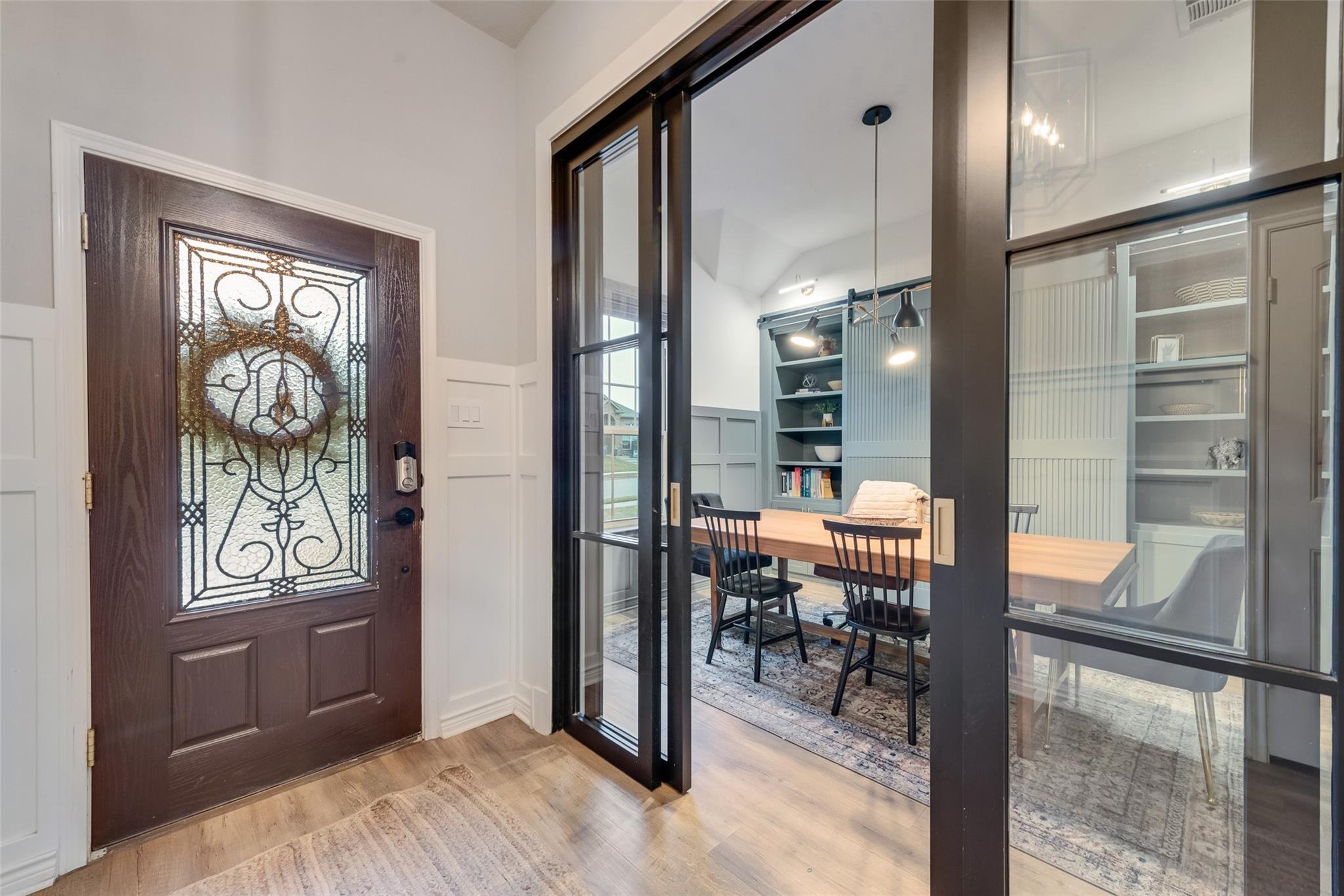 Interior view of an entryway featuring a dark brown wooden door with ornate glass. Adjacent is a glass sliding door leading to a modern office space with a wooden table, black chairs, built-in bookshelves, and pendant lights.