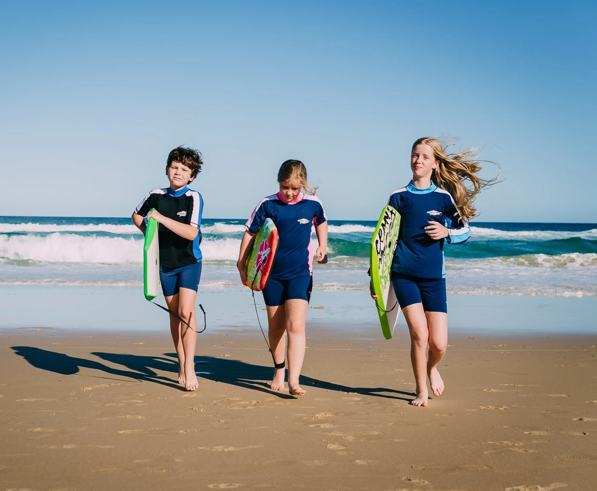 Three children walking on the beach with boogie boards, wearing matching navy and blue swimsuits.